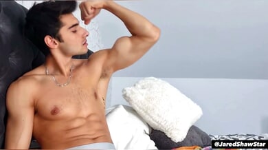 Romanian porn star Jared Shaw flexes his muscles and takes his big cock in his mouth on a gray tufted bed