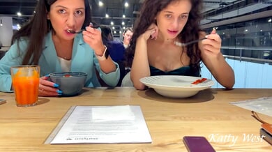 Katty West and a curly-haired woman dine bare beneath the table in a public restaurant